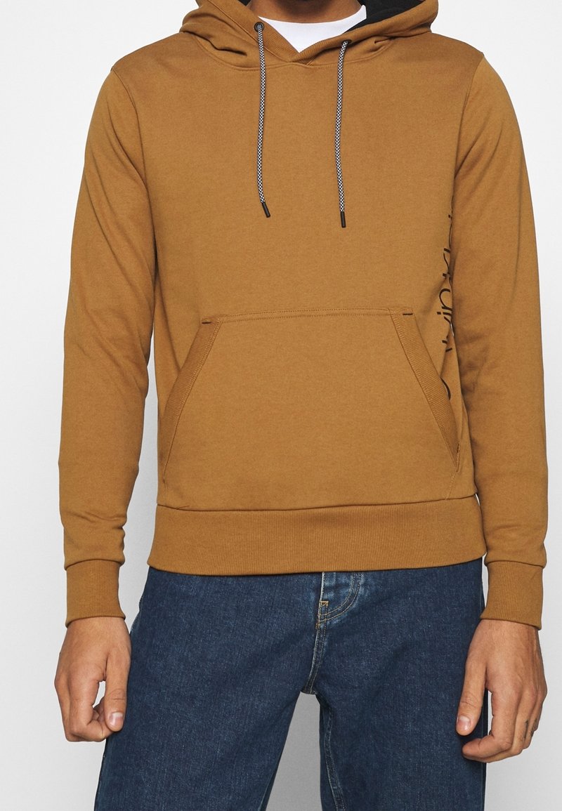 Sweat à capuche marron avec une poche kangourou, une capuche ajustable, et des cordons noirs. Présente une texture lisse ainsi que des poignets et un ourlet côtelés.