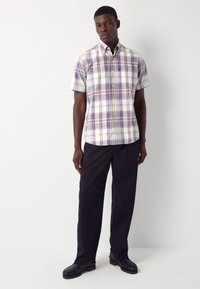 Mees, kes kannab lühikeste varrukatega lilla-valget ruudulist nööbitavat särki, musti pükse ja musti kingi, seisab valge tausta ees.