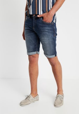 Shorts en denim bleu foncé avec ourlets retroussés, dotés de cinq poches et d'une fermeture éclair. Associés à des chaussures bateau gris clair.