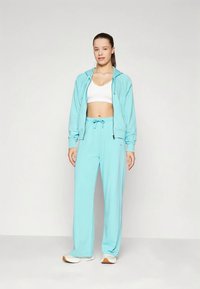 Žena stojí v svetlom modrom mikinovom saku, zodpovedajúcich teplákoch, bielom športovom podprsenke a bielych teniskách na jednoduchom pozadí.