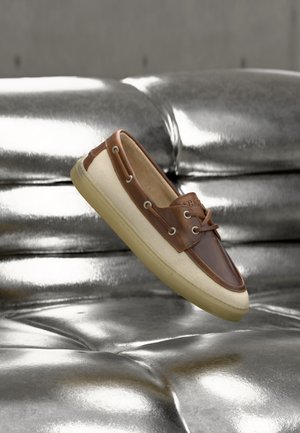 Zapato casual para hombre tipo slip-on con cuero marrón y tela beige, colocado sobre una superficie de cuero acolchada plateada brillante.