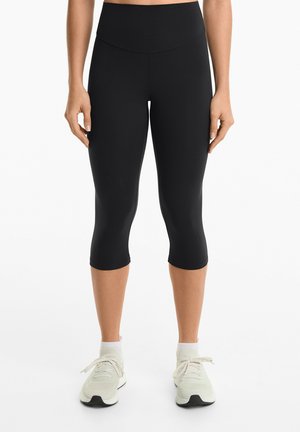 Leggings capri neri con vita alta, texture liscia e design aderente, abbinati a scarpe da ginnastica di colore chiaro.