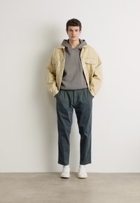Beige farmerdzseki egy szürke kapucnis felsőn, sötétzöld, tapered (szűkített) nadrággal és fehér sportcipővel párosítva. Egyszerű dizájn, minimális minták, laza megjelenés.