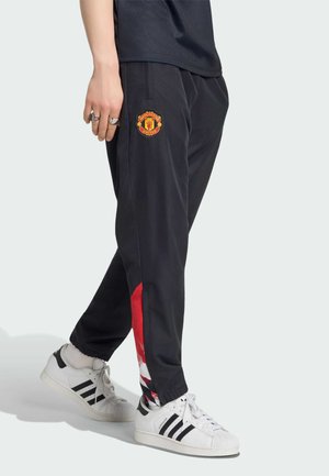 Persoon die zwarte Manchester United trainingsbroek draagt met rode en witte accenten en witte Adidas sneakers met zwarte strepen.