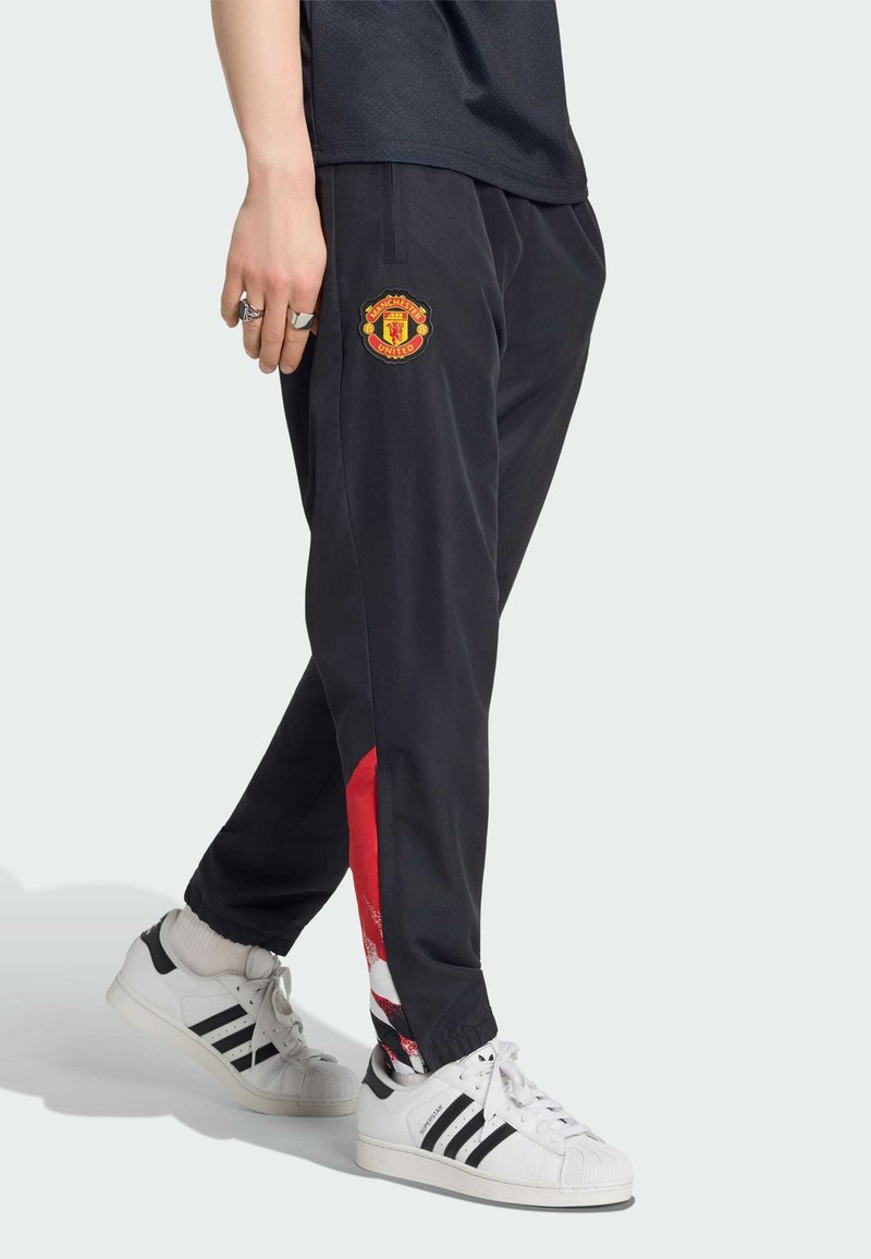 Personne portant un pantalon de survêtement noir Manchester United avec des accents rouges et blancs et des baskets blanches Adidas avec des bandes noires.