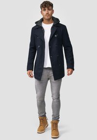 Marineblå peacoat med strukturert hette, dobbelspent knapper, kombinert med lysegrå jeans og brune snørebåndsstøvler.