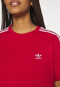 Žena s copánky oblečená v červeném tričku Adidas s bílými pruhy na ramenou a bílým logem Adidas na hrudi na bílém pozadí.