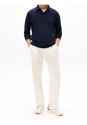 Marineblaues Langarm-Polo-Shirt mit Kragen, kombiniert mit weißen Hosen und beigen Sneakern. Sauberes Design mit minimalen Akzenten.