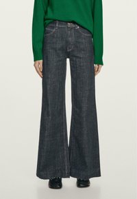 Jeans scuri a gamba larga con vita alta, caratterizzati da cuciture a contrasto e orlo grezzo. Abbinati a un maglione verde per creare contrasto.