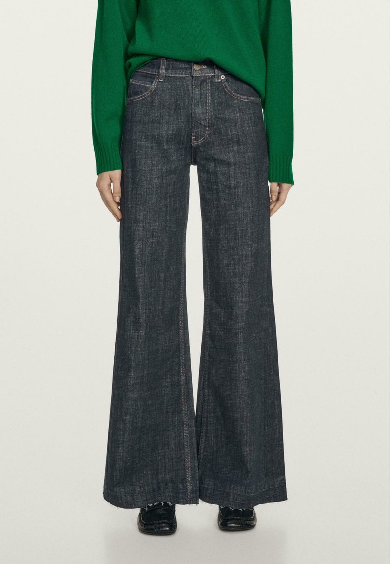 Jeans scuri a gamba larga con vita alta, caratterizzati da cuciture a contrasto e orlo grezzo. Abbinati a un maglione verde per creare contrasto.