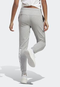 Grå träningsbyxor med tapered passform, elastisk midja och vitt logodetalj längs vänster ben. Bärs med vita sneakers.