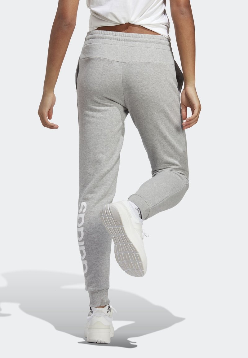 Grå träningsbyxor med tapered passform, elastisk midja och vitt logodetalj längs vänster ben. Bärs med vita sneakers.