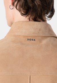 Hellbraune Wildlederjacke mit spitzem Kragen, die über eine sichtbare Stitching-Detailierung und ein silbernes "BOSS"-Logo auf dem Rücken verfügt. Glatte Textur.