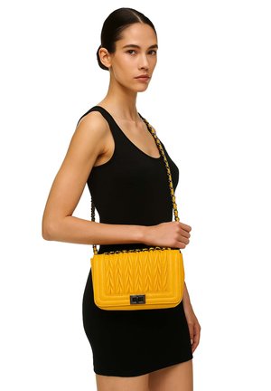 Donna in un abito nero senza maniche che tiene una borsa a tracolla gialla trapuntata con una tracolla a catena, rivolta verso sinistra su uno sfondo bianco.