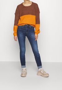 Kraftig stickad tröja med bruna och orange ränder, i kombination med smala blå jeans. Modellen bär beiga sneakers. Enkelt vit bakgrund.