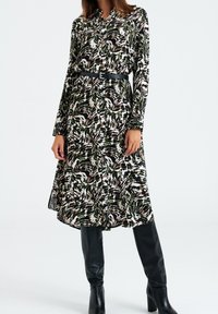 Abito camicia con un motivo astratto di foglie verdi, nere, bianche e marroni. Presenta maniche lunghe e una cintura nera in vita.