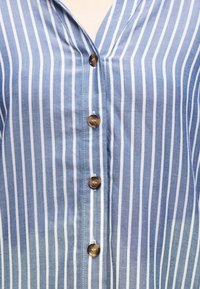 Πουκάμισο με κάθετες ρίγες σε μπλε και άσπρο, με καφέ κουμπιά που κουμπώνουν μπροστά, αποκαλύπτοντας το λαιμόκοψη σε σχήμα V.