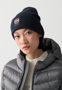 Sötétkék beanie, bordázott textúrával és logóval, szürke pufikabáttal és krém színű kötött pulóverrel párosítva. Arcközeli felvétel.