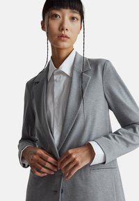 Šedý blazer s klopami a dvěma knoflíky, který má hladkou texturu. Spárovaný s bílou košilí s knoflíky pod ním.