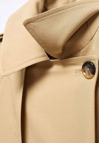 Col de trench-coat beige avec des coutures détaillées et un bouton en écaille foncée fixé près de l'encolure.