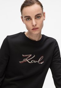 Femme avec les cheveux plaqués en arrière portant un sweat à capuche noir avec "Karl" en sequins sur le devant, sur fond blanc uni.