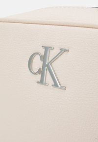 Calvin Kleinin käsilaukku pehmeästä beige-kuvioidusta materiaalista, jossa on hopeanvärinen "CK"-logo. Selkeä, moderni muotoilu ja jämäkkä rakenne.