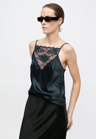 Kobieta z gładko zaczesanymi włosami, nosząca czarne prostokątne okulary przeciwsłoneczne, czarny satynowy top na ramiączkach z kwiatową koronką oraz pasującą czarną spódnicę.
