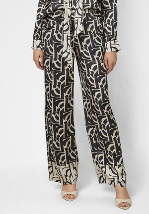 Frau trägt schwarze und beige weit geschnittene Hosen mit geometrischem Muster, dazu passendes Oberteil und beige offene High Heels, steht vor weißem Hintergrund.