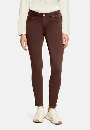 Femme portant un jean slim marron, un haut blanc à manches longues et des baskets blanches, debout devant un fond clair uni.