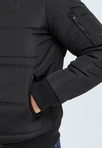 Black padded jacket with ribbed cuffs, featuring side and arm zippers. The material is smooth, with a lightweight, insulated design.