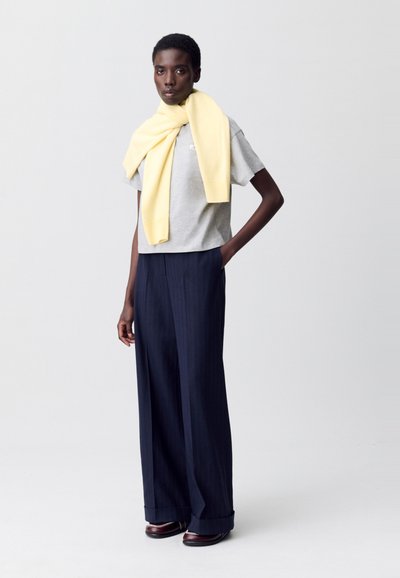 Camicetta grigia a maniche corte, sciarpa di lana gialla appoggiata sulle spalle, pantaloni a gamba larga blu navy con motivo a righe, scarpe marroni con dettagli metallici.