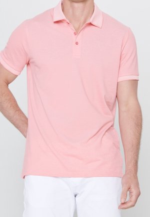 Mann trägt ein hellrosa kurzärmeliges Poloshirt mit Kragen und drei Knöpfen, kombiniert mit weißen Hosen.