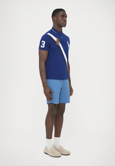 Tengerészkék galléros póló fehér átlós csíkkal és hímzett logóval, világoskék rövidnadrággal és bézs mokaszin cipővel, fehér zoknival párosítva.