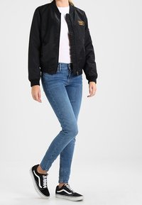 Chaqueta bomber negra con cremallera frontal, adornada con un parche de logo dorado, combinada con jeans ajustados azules y zapatillas Vans negras.