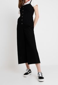 Svart denim jumpsuit med vida ben, knapprad framtill och justerbara axelremmar. Bärs med en vit skjorta och svarta sneakers.