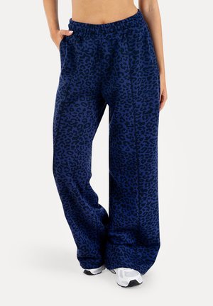 Kobieta w luźnych granatowych spodniach w panterkę z elastycznym pasem i białych trampkach, stoi z jedną ręką w kieszeni.