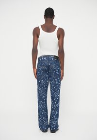 Vyras, vilkintis baltą be rankovių marškinėlį ir mėlynus raštuotus džinsus, stovi nugara į priekį prieš plokščią foną.