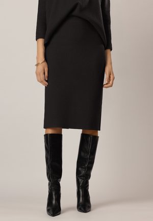 Femme portant une jupe noire longueur genou, un haut à manches longues noir, un bracelet doré et des bottes en cuir noires montant jusqu'aux genoux sur un fond uni.