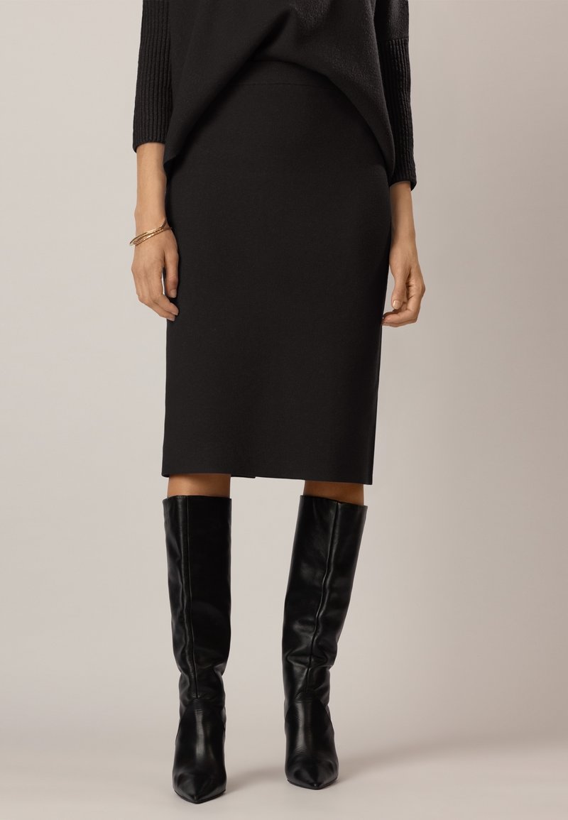 Femme portant une jupe noire longueur genou, un haut à manches longues noir, un bracelet doré et des bottes en cuir noires montant jusqu'aux genoux sur un fond uni.