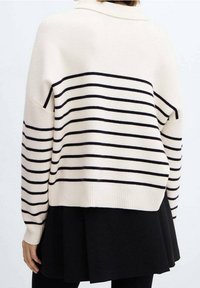 Pull crème avec des rayures marines, présentant un col et des poignets côtelés. L'arrière a une coupe ample et l'apparence d'une jupe noire superposée.