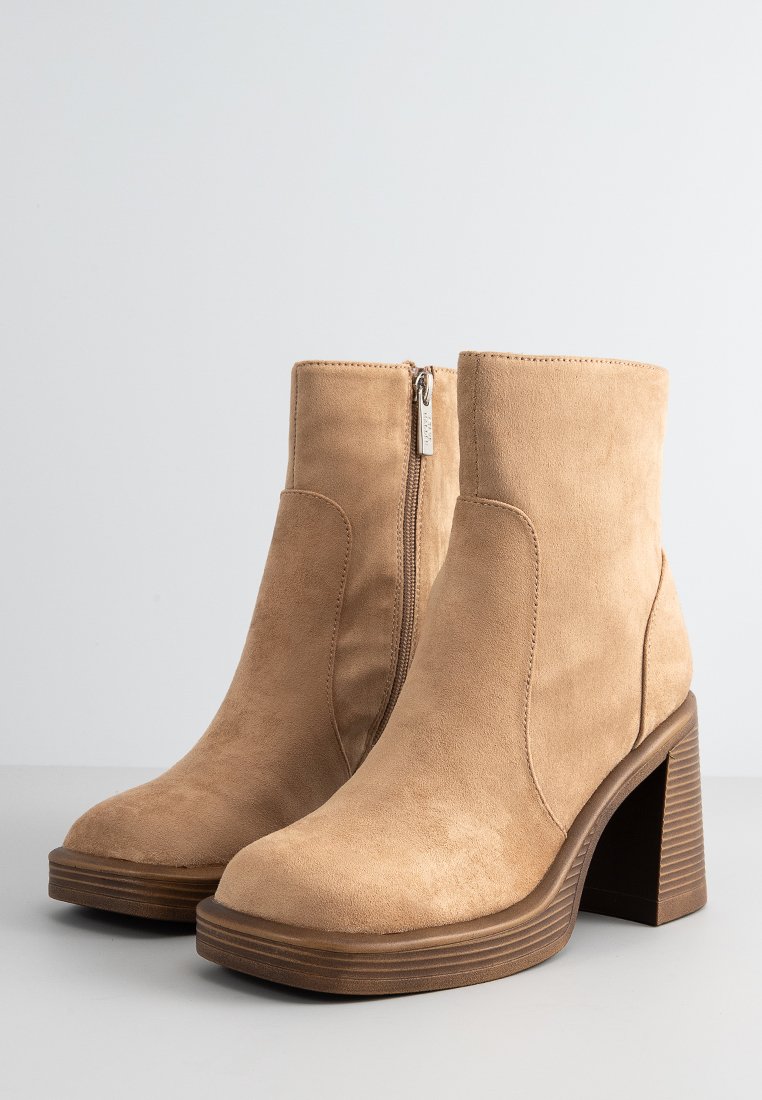 Bottines en suédine beige avec une fermeture éclair sur le côté, talon épais brun, embout arrondi et texture de semelle côtelée.