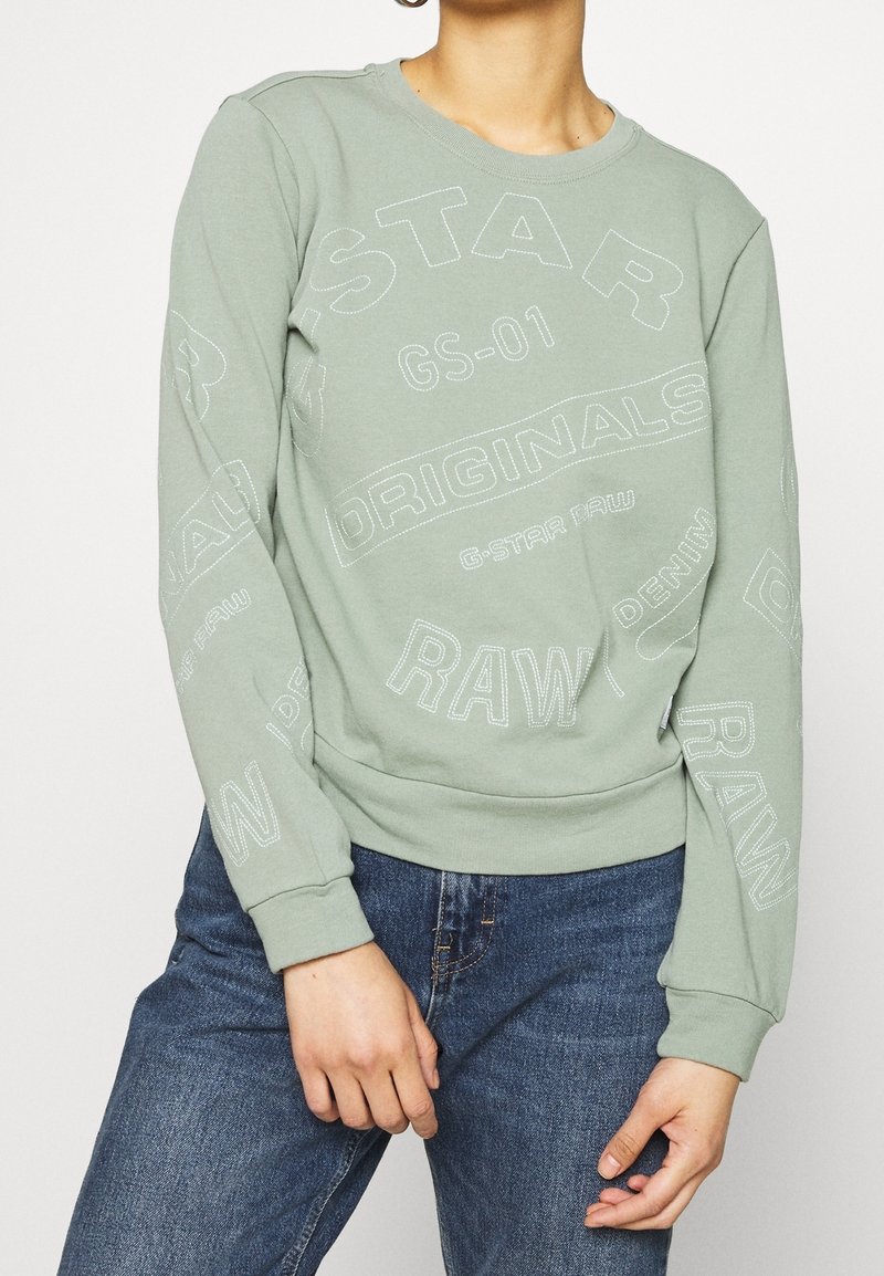 Sudadera verde claro con cuello redondo, con gráficos y texto en blanco en negrita. Fabricada en un tejido suave, con mangas largas y una cintura ajustada.