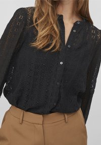 Czarna koronkowa bluzka z wysokim dekoltem, zapięciem na guziki i długimi rękawami. Posiada skomplikowane wzory oraz teksturowany materiał w połączeniu z beżowymi spodniami.