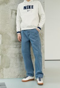 Vit hoodie med marinblå "NIKE"-logotyp, blå jeans och vita sneakers med gummisulor, mot en betongbakgrund.