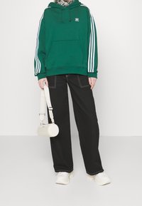 Zelená mikina s bílými pruhy, přední kapsou a logem; kombinovaná s černými širokými kalhotami a bílou válcovitou taškou, doplněná robustními bílými teniskami.