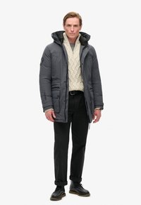 Veste en duvet grise avec une capuche, portant un pull texturé crème en dessous, un pantalon noir et des chaussures à lacets noires.