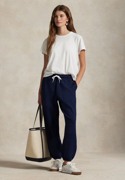 Γυναίκα που φοράει λευκό t-shirt, σκούρο μπλε παντελόνι φόρμας, λευκά αθλητικά παπούτσια, κρατώντας μπεζ και μαύρη τσάντα tote, στέκεται μπροστά σε ουδέτερο φόντο.