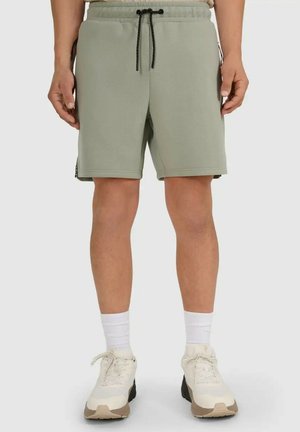 Mężczyzna w jasnozielonych szortach ze sznurkiem, białych skarpetkach oraz białych butach sportowych, stojący na jednolitym tle.