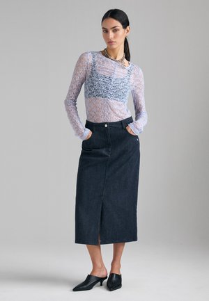 Donna che indossa un top in pizzo trasparente a manica lunga con canotta corta, gonna midi scura a vita alta, mule nere e collana a catena, mani in tasca.