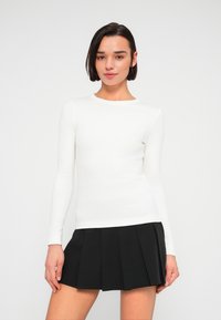 Λευκή μακρυμάνικη εφαρμοστή μπλούζα με ριμπ ύφασμα, συνδυασμένη με μαύρη πλισέ φούστα. Η λαιμόκοψη είναι στρογγυλή και η εφαρμογή στενή.